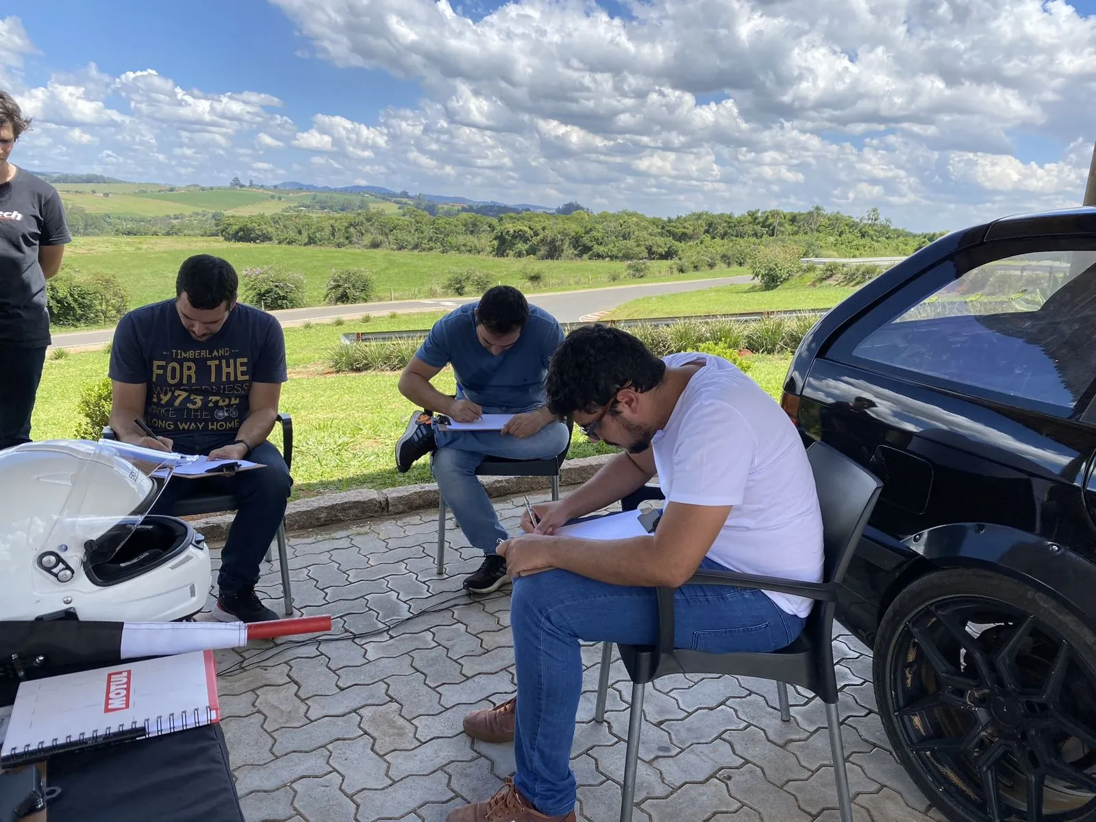 Alunos fazendo anotações em pranchetas durante o briefing, com a pista ao fundo