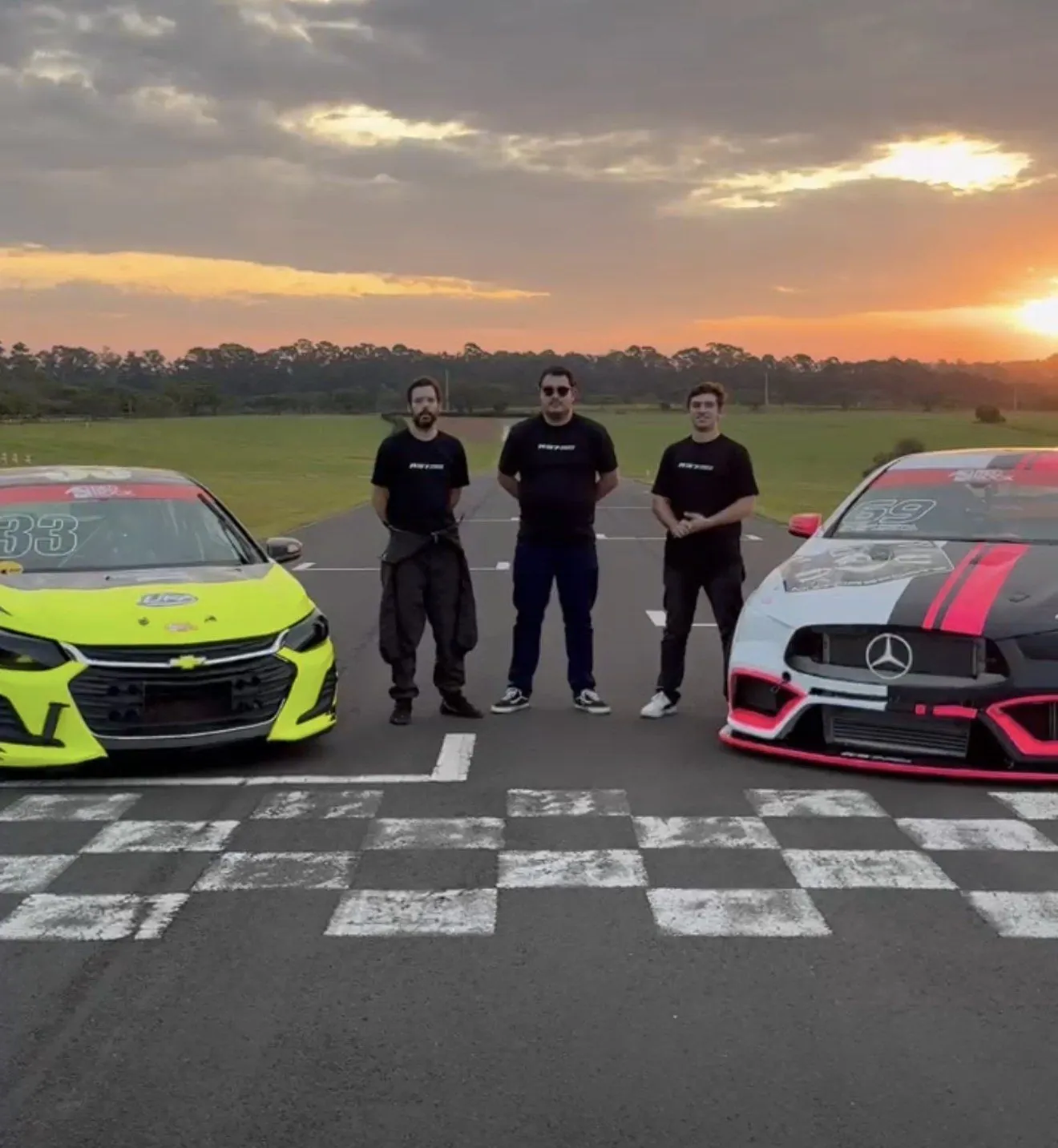 Os três sócios da R37 Driver Academy na linha de largada, entre dois carros de corrida, ao pôr do sol, durante um evento corporativo