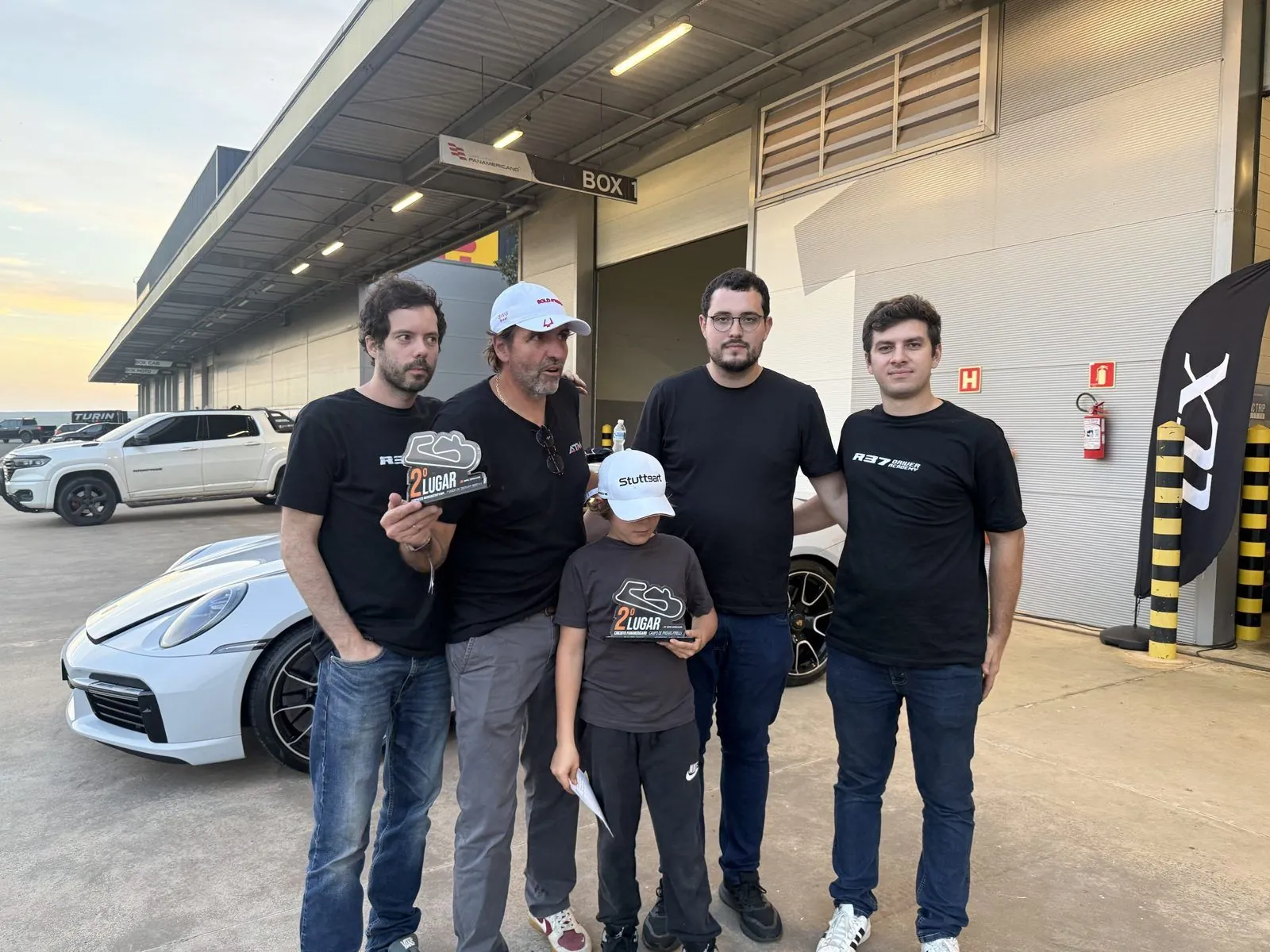 Equipe da R37 Driver Academy ao lado de um cliente com o troféu de 2º lugar, em frente aos boxes do Circuito Panamericano