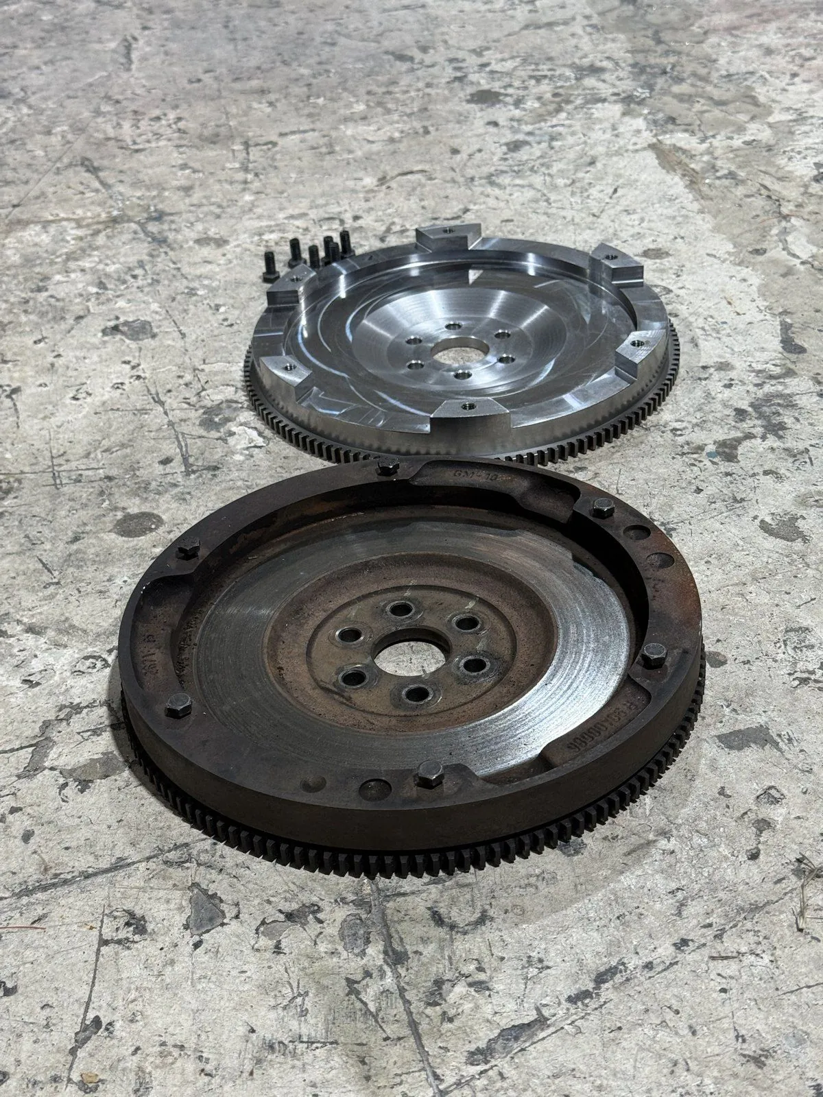 Volante em aço billet usinado, com os parafusos novos, ao lado do volante original do motor GM, sobre o chão da oficina