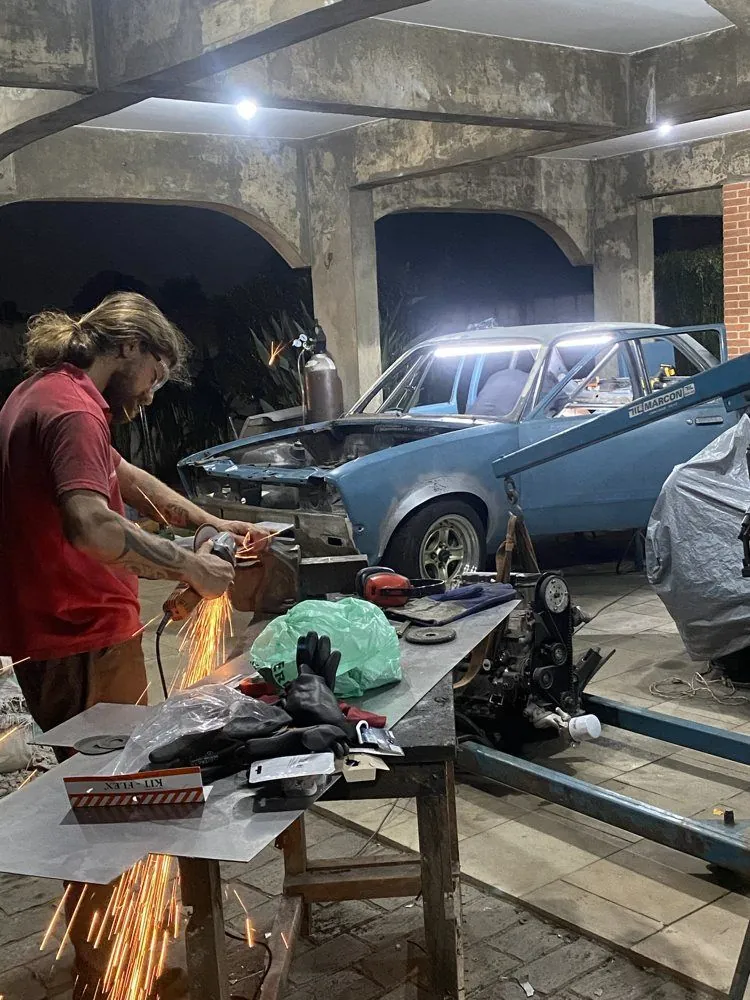 Amigo cortando chapa com esmerilhadeira, com o CHEV37TE ao fundo na oficina improvisada
