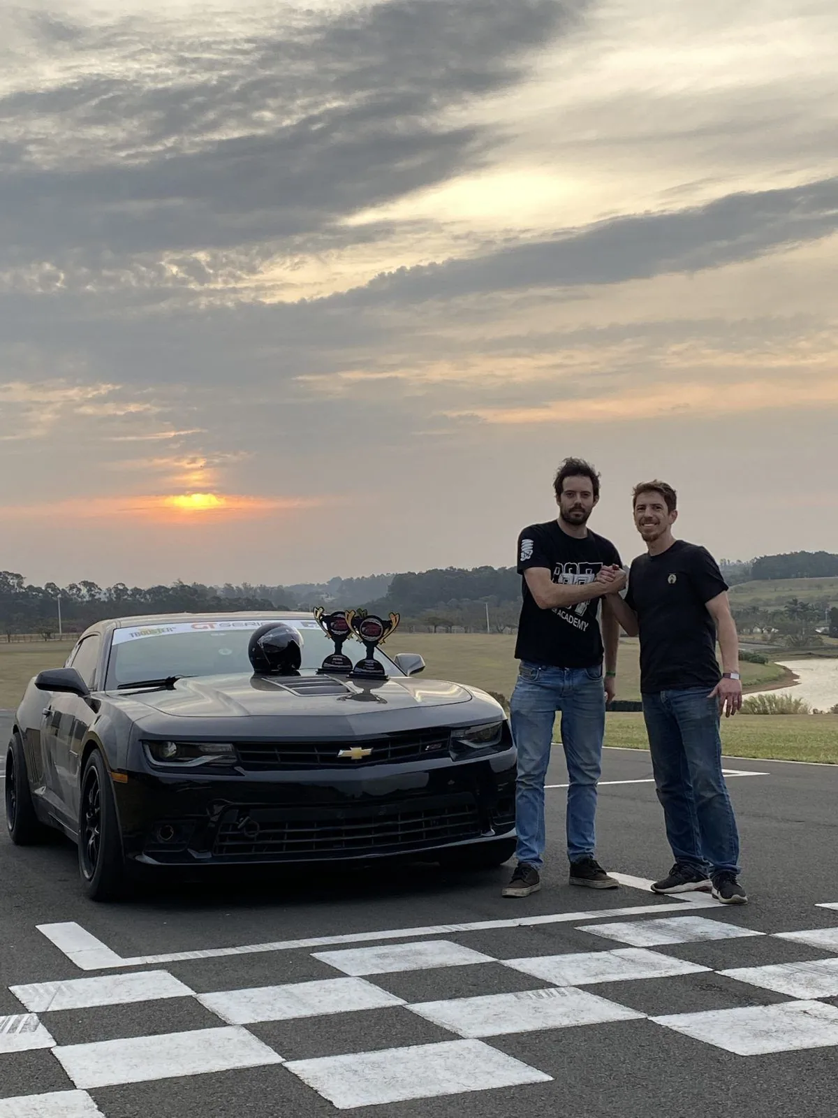 Ricardo Gouveia e Rodrigo Mourão se cumprimentam ao lado do Camaro preto, com dois troféus sobre o capô, ao pôr do sol