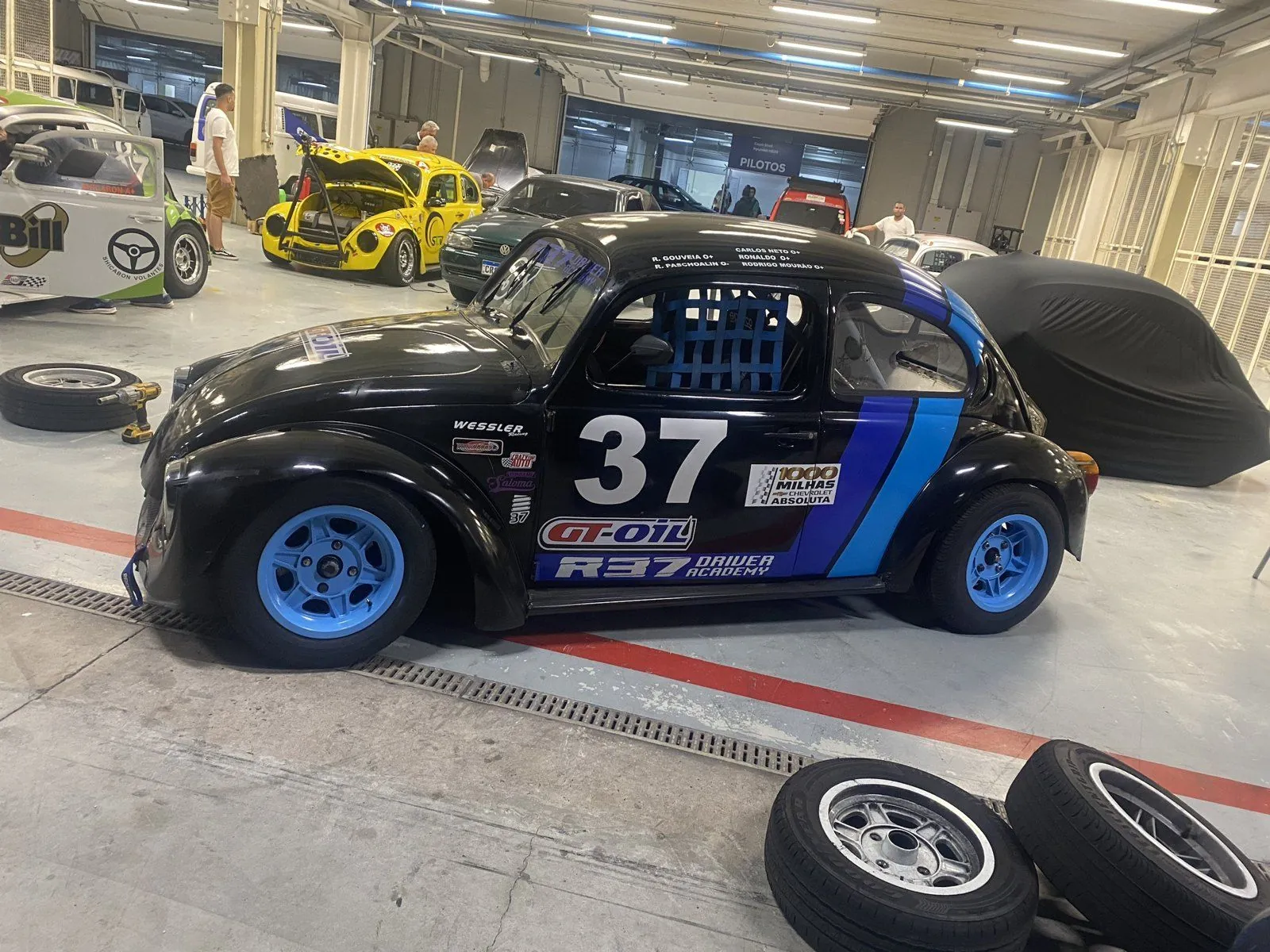 Fusca 37 de lado no box, com rodas azuis e outros Fuscas de corrida ao fundo
