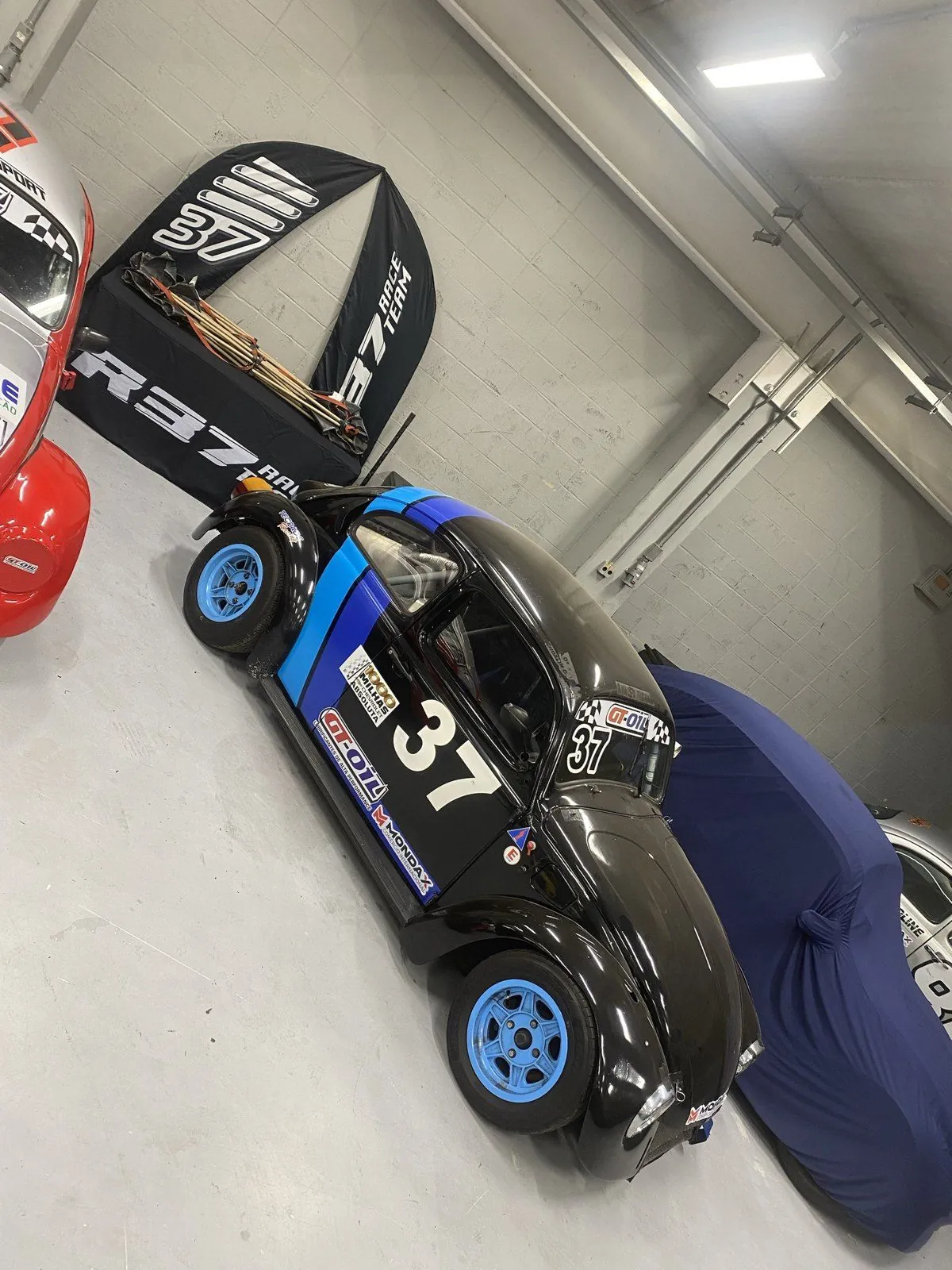 Fusca 37 preto com faixas azuis e rodas azuis nos boxes, ao lado da mesa e das bandeiras da R37