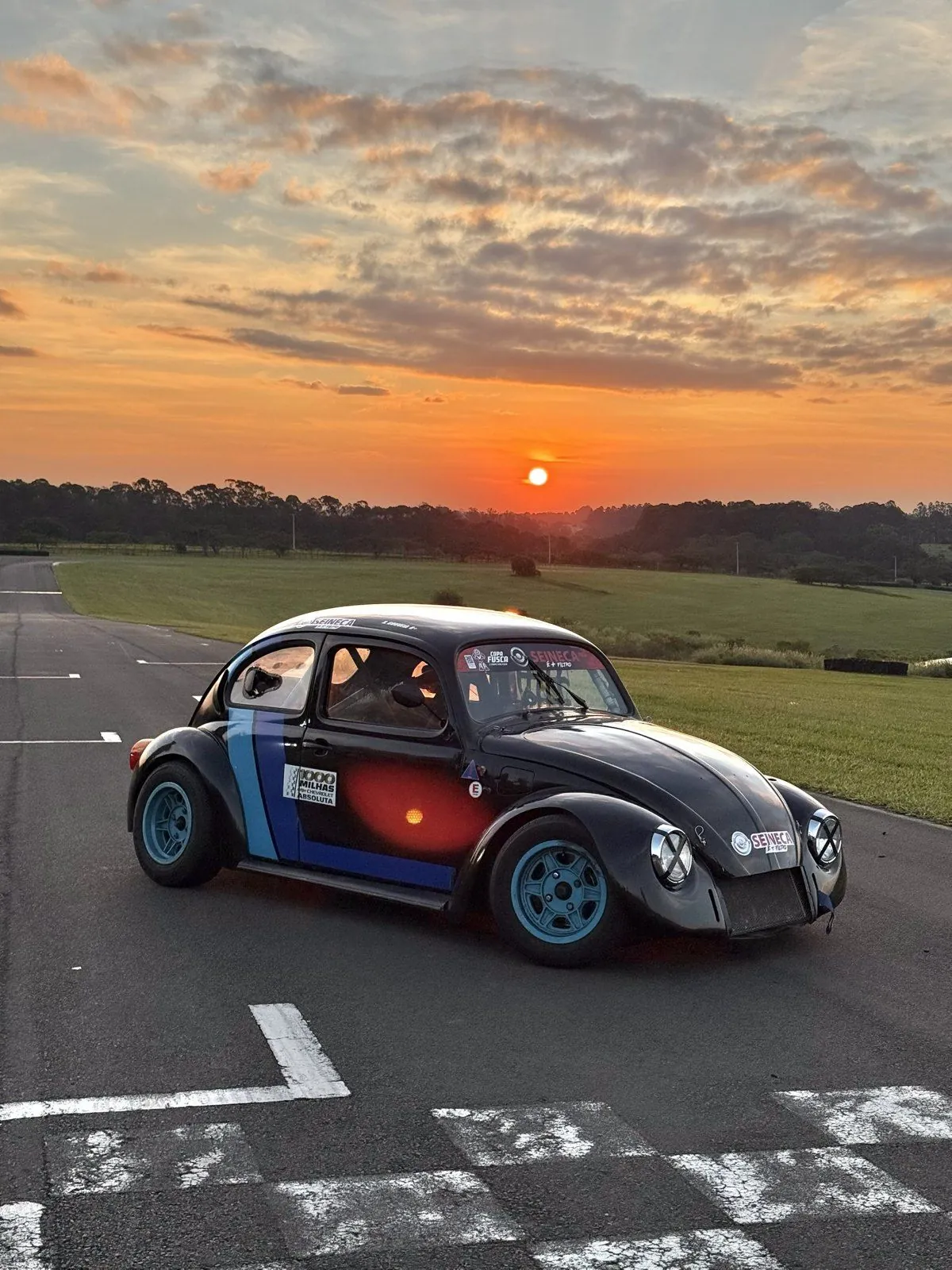 Fusca 37 preto com faixas azuis parado na pista, com o sol se pondo atrás