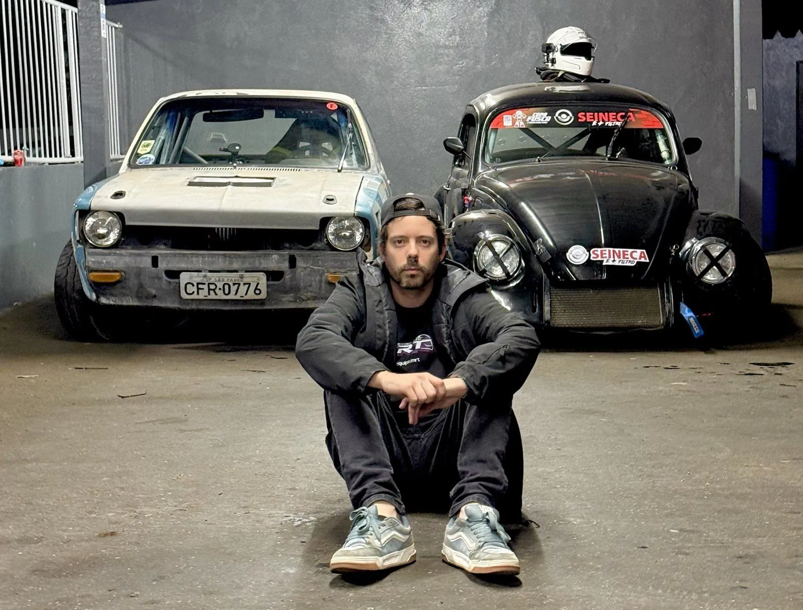 Ricardo sentado no chão do box, à noite, com o CHEV37TE e o Fusca 37 estacionados lado a lado atrás dele