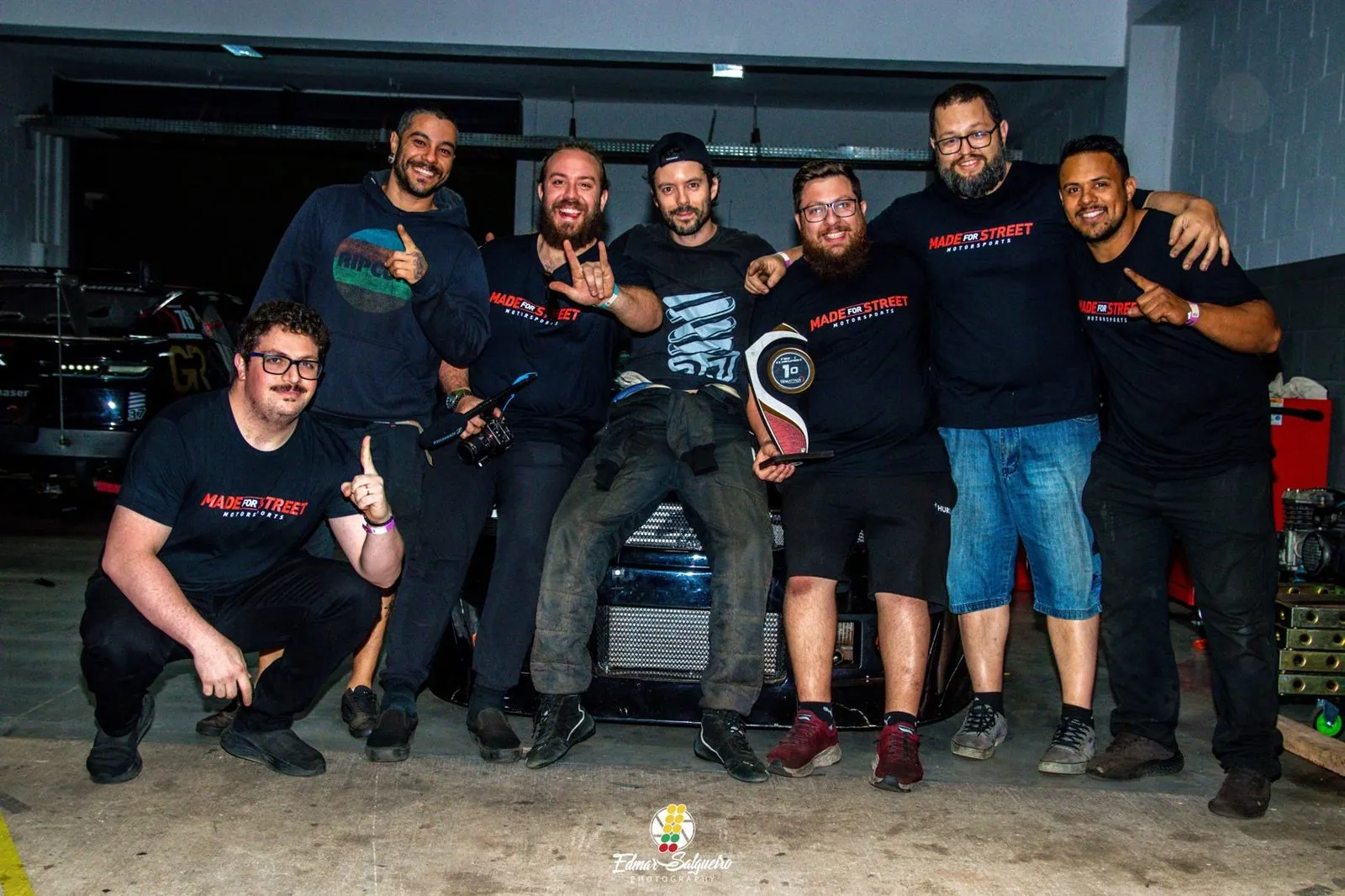 Ricardo sentado na frente do carro, com a equipe Made for Street e o troféu de 1º lugar, no box