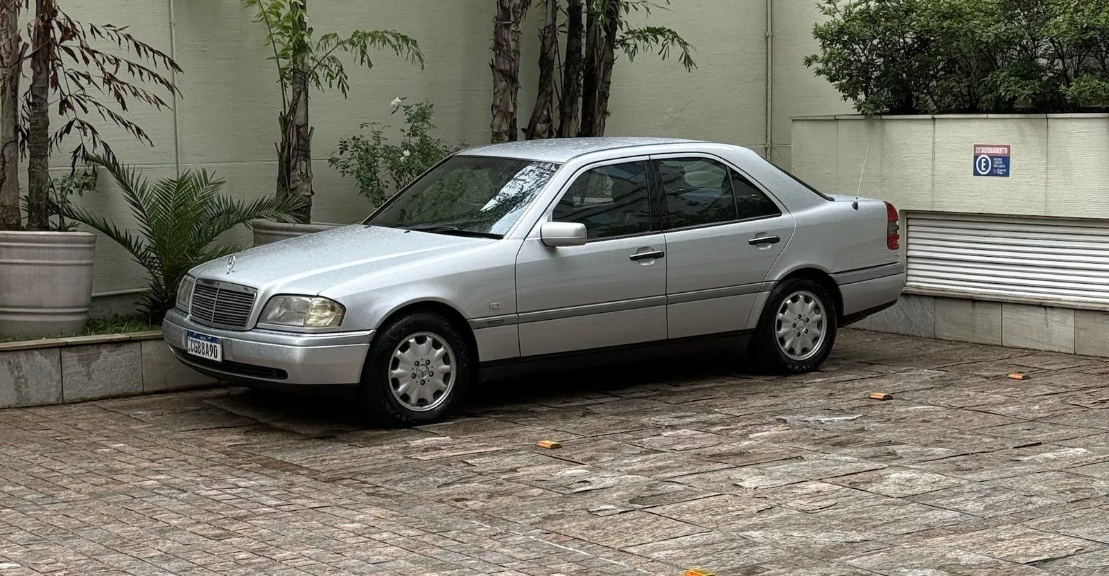Mercedes C280 1996 prata estacionada num pátio de pedras, com plantas ao fundo