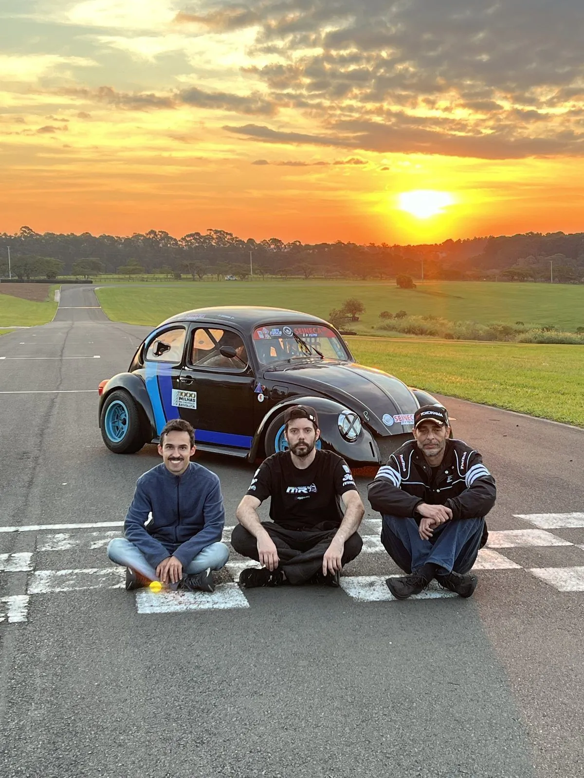 Ricardo, Daniel Pontes e Lucas Ferreira sentados na linha de chegada da Fazenda Capuava ao pôr do sol, com o Fusca de Copa Fusca ao fundo